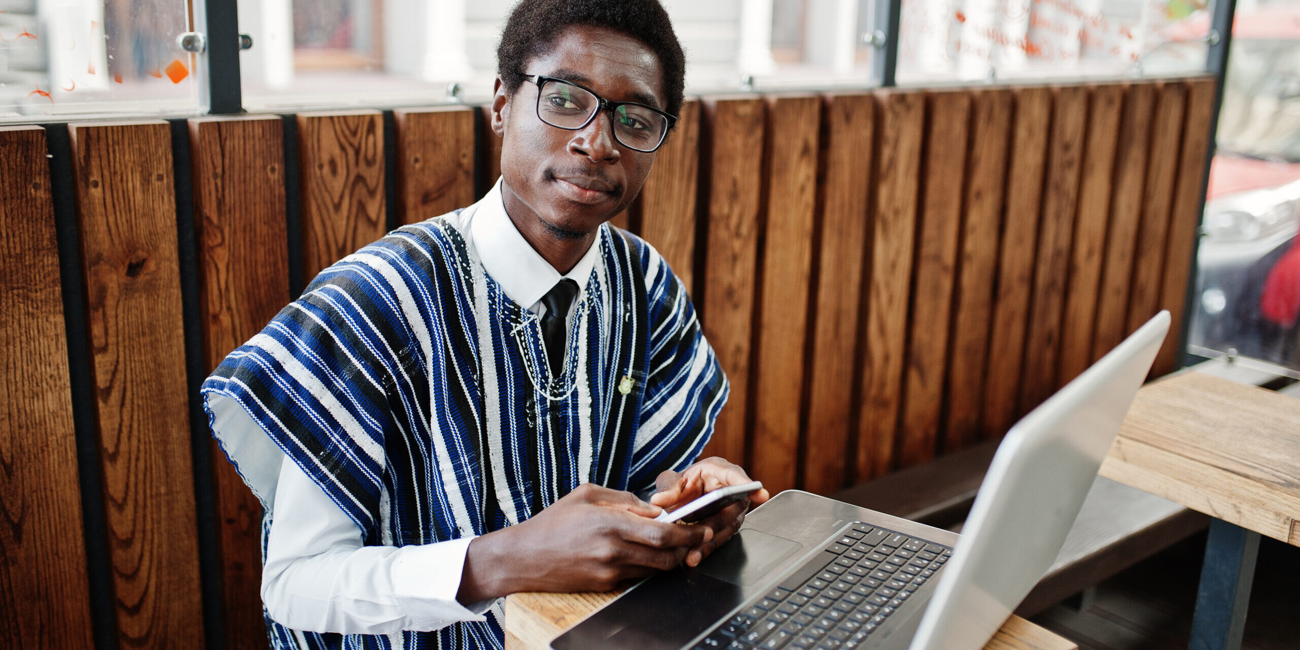 African man in traditional clothes and glasses sitting behind laptop at outdoor caffe and looking on mobile phone.
