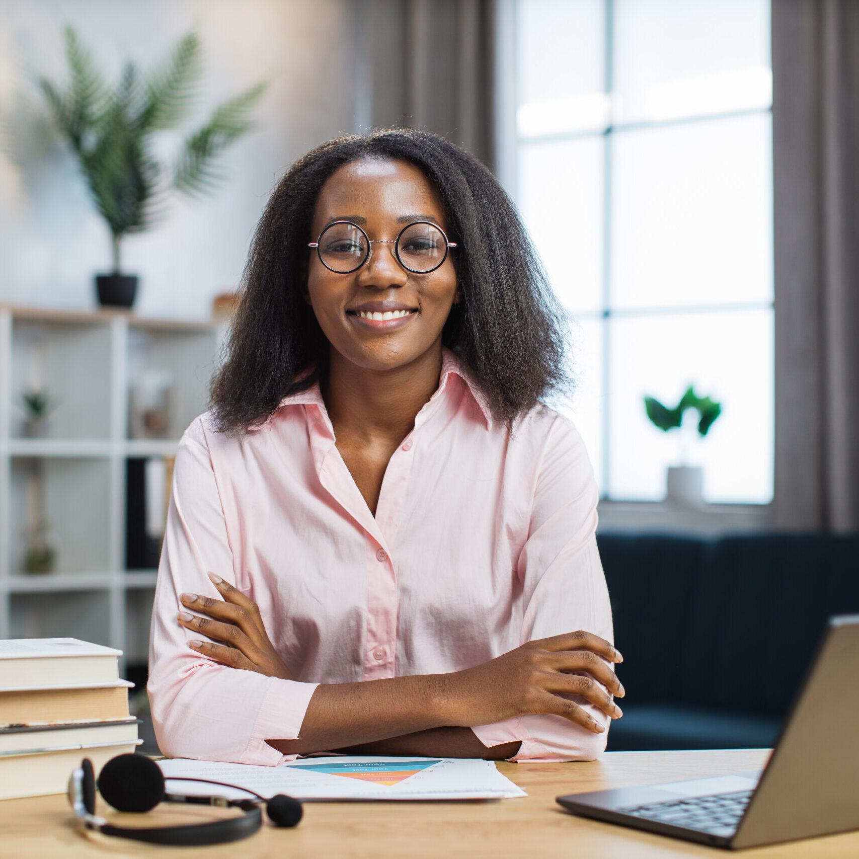 Smiling african american woman in eyeglasses and pink shirt sitting at desk with books and notes. Female tutor working at home during distance learning.