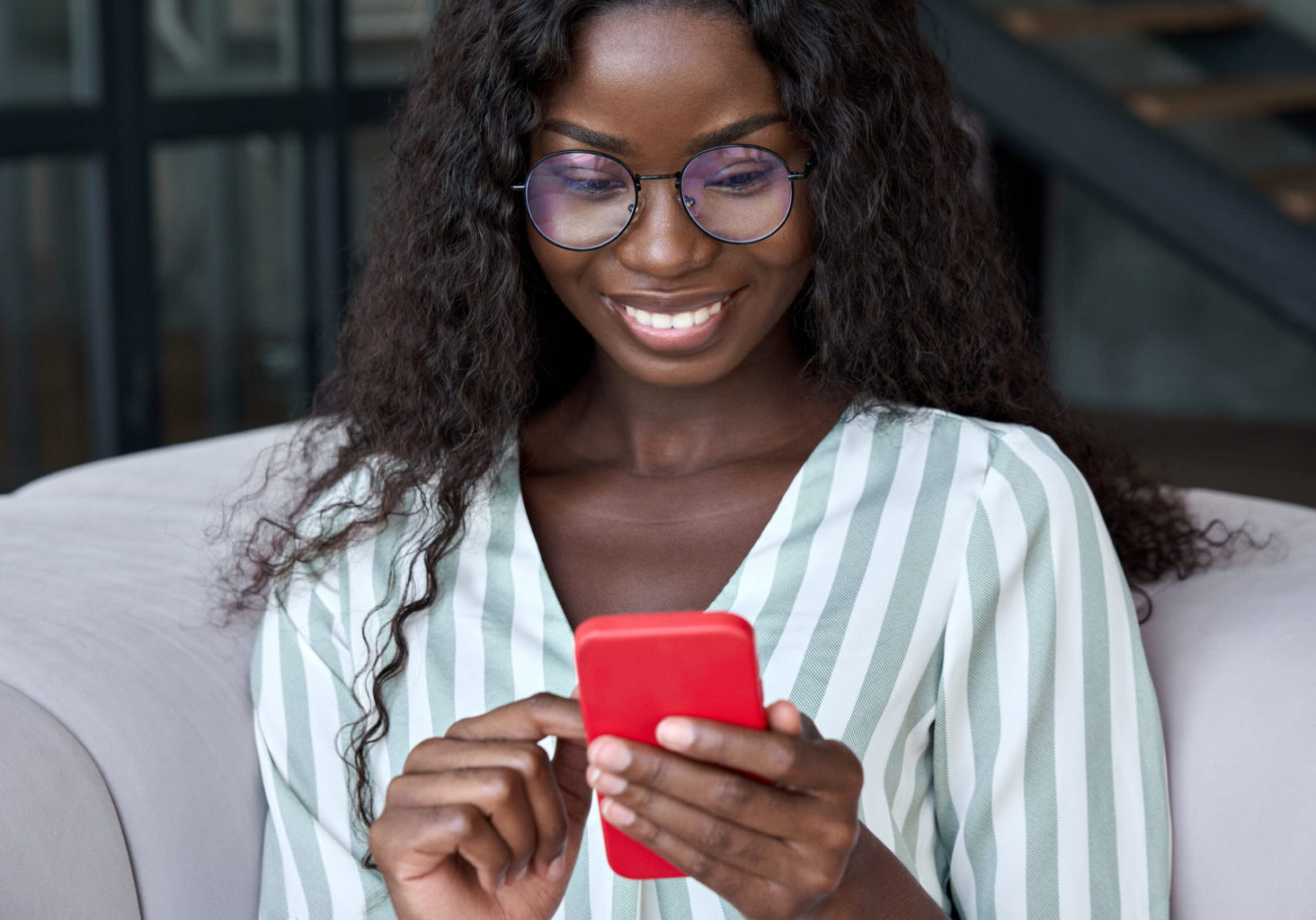 Young happy black African woman wearing glasses using mobile digital apps on cell phone tech device for online shopping or dating sitting on sofa at home, texting on smartphone, writing social post.