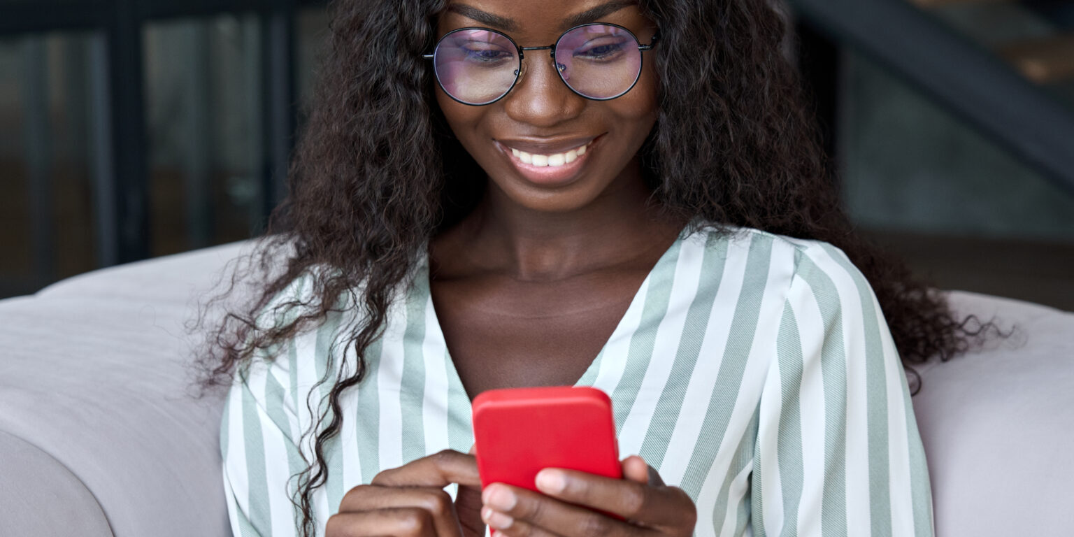 Young happy black African woman wearing glasses using mobile digital apps on cell phone tech device for online shopping or dating sitting on sofa at home, texting on smartphone, writing social post.
